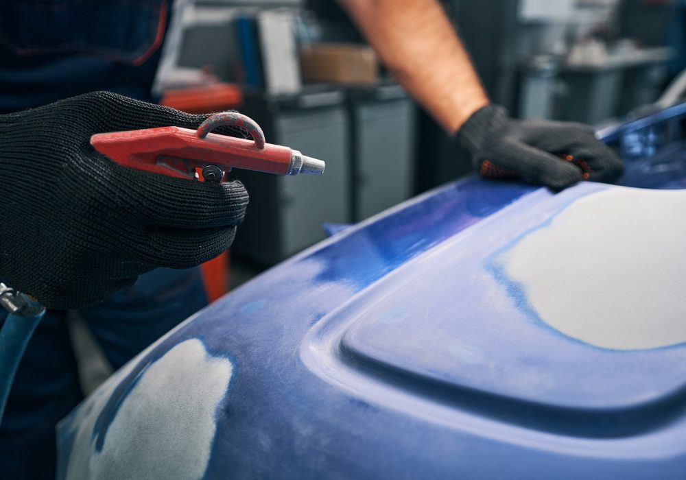 Worker blowing air on blue metal car body part from instrument after sanding paint from its surface